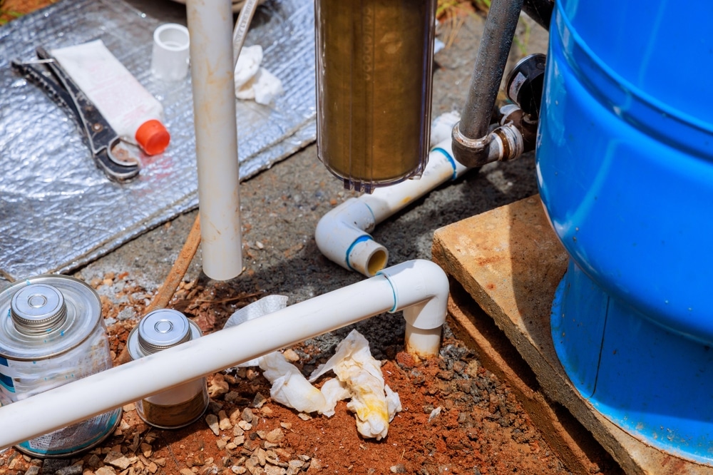 Maintenance de système de puits et plomberie extérieure Réparation d'un système de puits avec tuyaux PVC, réservoir bleu, filtre à sédiments et outils de plomberie au sol.